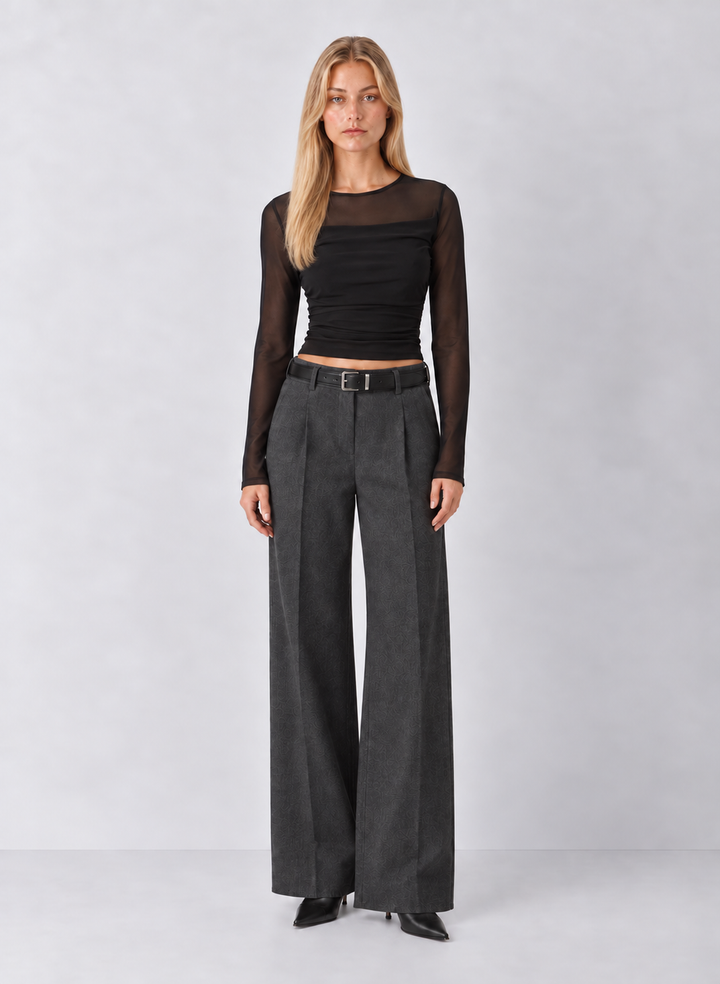 A woman stands against a light gray studio background wearing a black sheer long-sleeve cropped top and dark charcoal wide-leg trousers with a thin black belt.